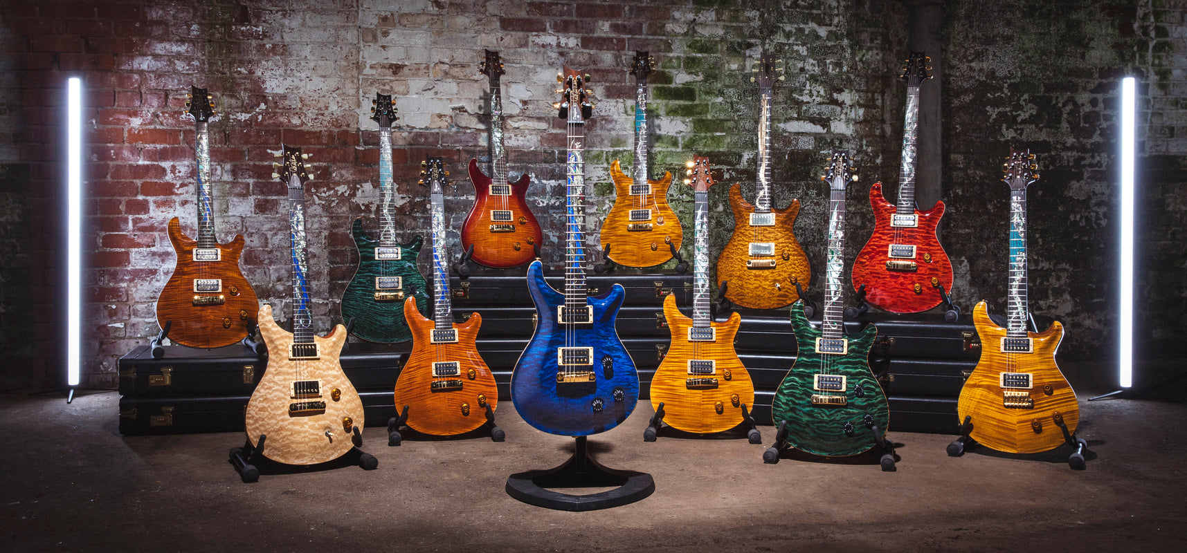 A collection of PRS guitars in various colors: blue, green, and shades of brown, with quilted maple tops and unique finishes, displayed on stands.
